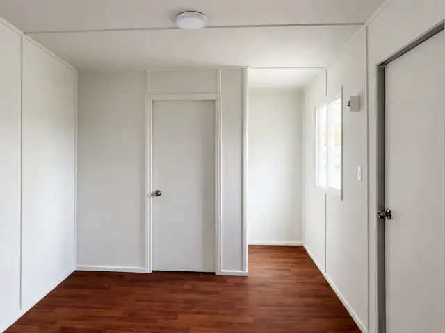 Tabique del medio baño con su puerta, junto a la puerta de acceso, sobre muros forrados y piso vinílico oscuro