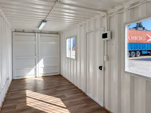 Interior sin forrar en planta libre: el corrugado del contenedor pintado de blanco, piso vinílico símil madera y las puertas originales al fondo