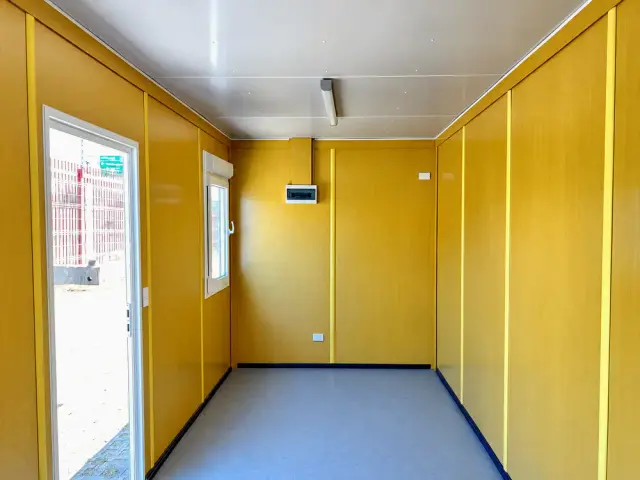 Interior de la oficina de 20 pies sin divisiones: paneles amarillos, piso vinílico gris, tablero eléctrico y tubos fluorescentes